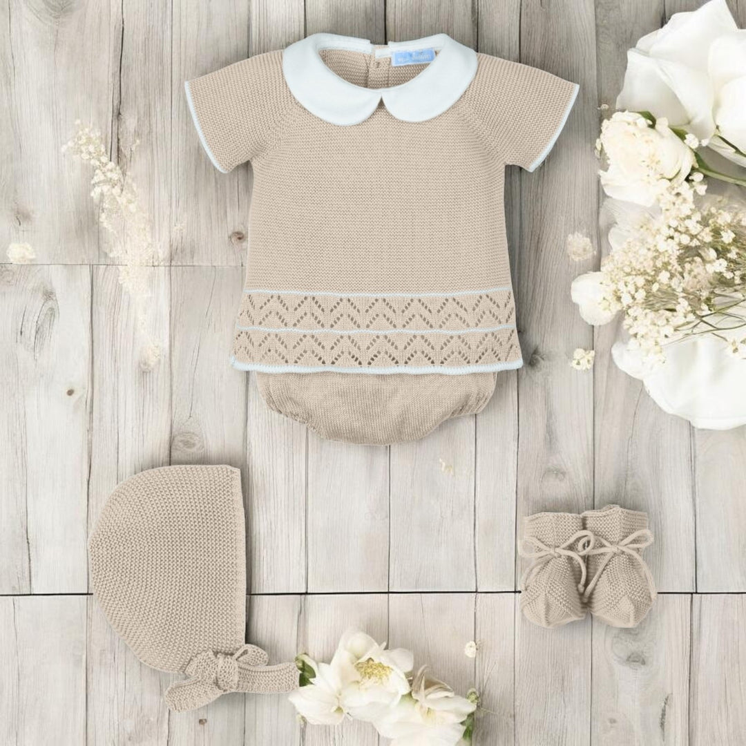 Baby outfit with top, bloomers, bonnet, and shoes on a wooden surface with flowers.