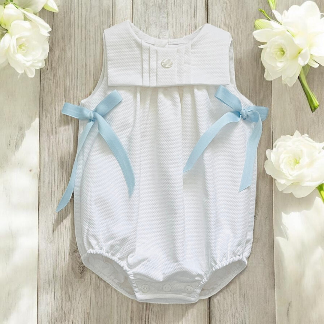 White baby romper with blue bows on a wooden surface with white flowers.