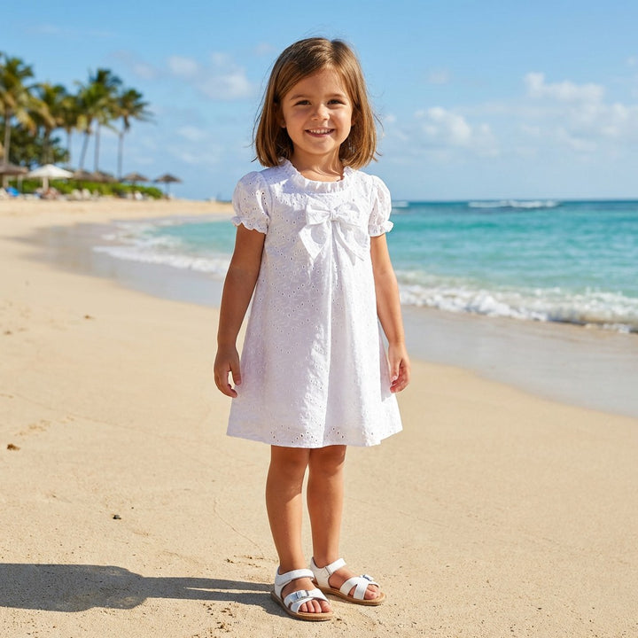Young girl in a white dress standing on a sandy beach with ocean and palm trees in the background.